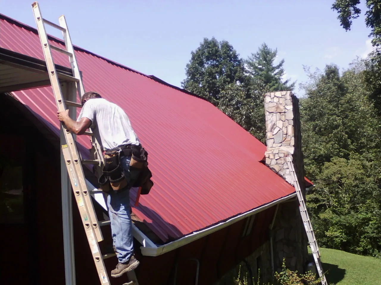Roofer on a ladder beside a red metal roof and stone chimney — Soffit & Fascia Repair by Roofing Brothers in Ithaca